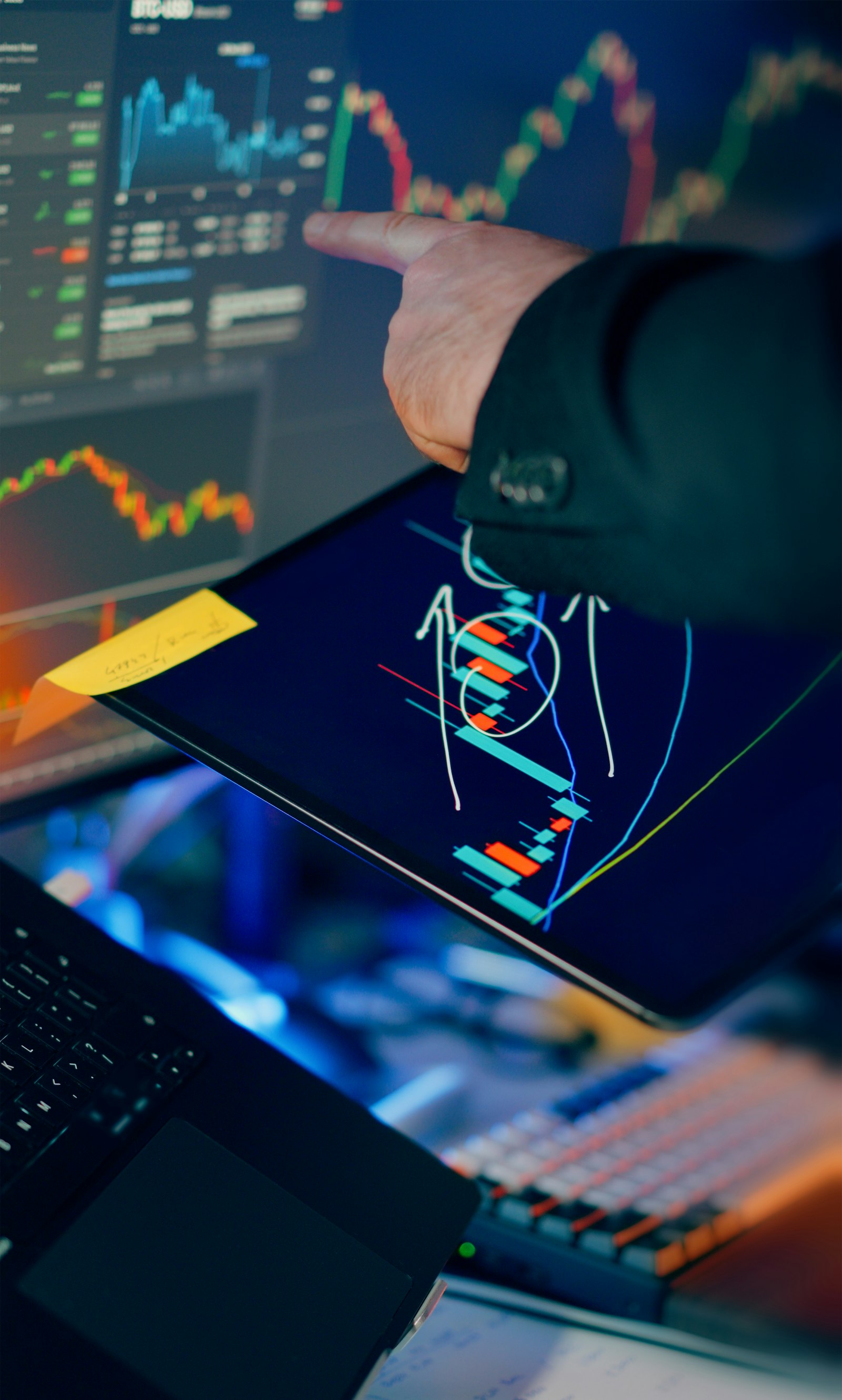 A trader's hand pointing at a touchscreen showing candlestick charts, with multiple monitors, a laptop and a tablet in view